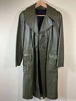 Front view of dark olive green leather coat hanging, showing double-breasted snap buttons, large lapel collar, flap pockets