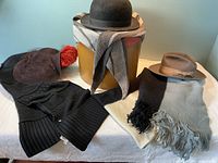 Photo shows three vintage wool hats: a black bowler, a brown fedora, and a ladies hat with black net and cranberry feather, arranged around a tall vintage hat box. Three wool scarves displayed: a long black knit UGG scarf, a grey striped thin triangle-shaped scarf, and a thick, long, wide shawl in black, cream, and black stripes.