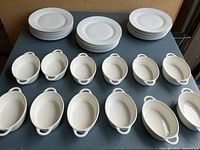 Top view of 24 white round plates arranged in three stacks behind 12 oval two-handled au gratin dishes lined up in two rows on a table.