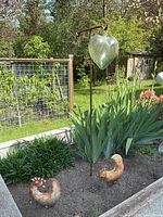 Glass heart ornament suspended from tall metal stake in garden bed with plants, two rusted metal bird sculptures at base.