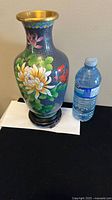 Front view of the blue Chinese cloisonné vase on wooden stand with a water bottle for size reference.