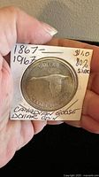 Front side showing Canadian Goose soaring with text 'DOLLAR' and 'CANADA 1867-1967'. Coin is in a paper holder with handwritten notes.