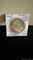 Front side of 1965 Canadian Voyager silver dollar coin held in white coin holder with handwritten notes.