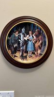 Front view of the Wizard of Oz collector plate mounted in a wooden round frame, showing main characters Dorothy, Scarecrow, Tin Man, Cowardly Lion, and dog Toto on yellow brick road.
