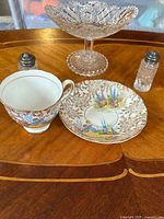 Five-piece lot arranged on a wooden surface including the patterned cup and saucer, salt and pepper shakers, and vintage glass candy dish.