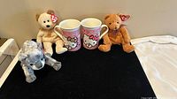 Two Hello Kitty pink coffee mugs and three Ty Beanie Babies plush bears arranged on black surface.