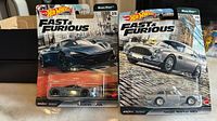 Front view of two Hot Wheels Fast & Furious cars, McLaren 720S in black and Aston Martin DB5 in silver, still sealed in original packaging.