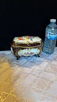 Closed trinket box beside water bottle to show size and detail of floral and gold accent decorations on footed porcelain box.