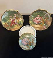 Photo of two hand-painted porcelain plates with romantic pastoral scenes and a matching round trinket box lid depicting a seated woman holding fruit. The items rest on a black surface with plate stands.