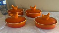 Four vintage orange mini casserole dishes with lids, chicken-shaped handles, set on a white cloth surface.