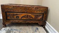 Front view of the rectangular wooden trunk featuring detailed hand carved floral, geometric, and scenic decorations. It rests on four carved claw feet.