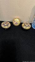 Front-facing view of three cloisonné trinket boxes on black fabric, showing colorful floral enamel designs on gold backgrounds.