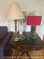 Four table lamps placed on a wooden table next to a couch, including tall brass lamp with beige shade, red shade lamp, clear glass lamp, and white base lamp.