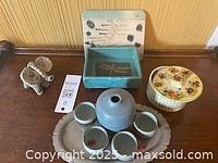 Photo shows the lot arranged on a wooden surface against a yellow wall. Items include a blue saki set with bottle and cups on an oval tray, a small elephant figurine, a blue box, and a floral trinket box.