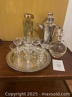 Front view of vintage silverplate cocktail shaker, nine cocktail glasses arranged on silverplate tray, and a glass decanter with stopper on a wooden surface against a wood panel background.