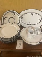 Grouped photo showing stack of 7 plates, 3 platters behind them, 2 coffee cups on saucers in front on wooden surface.