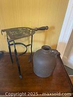 Vintage brass fireplace trivet with ornate cut-out design and handle, positioned next to old dark brown jug on wooden surface.