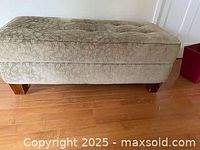 Heavy upholstered ottoman with tufted button top and wooden legs, positioned on hardwood floor against wall.