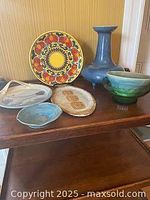 Photo of vintage pottery and glassware collection on wooden shelf including colorful plate, blue vase, hand-thrown bowl, glass bowl, and platter.