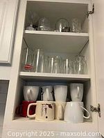 Upper shelf of a cabinet displaying clear glass drinking glasses including some with embossed designs, and lower shelves with a red mug, cream mug with leaf design, and several white mugs.