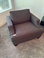 Brown fabric armchair by Van Gogh Designs shown in natural light next to a window and carpeted floor, with visible cushioned seat and backrest, and padded armrests.