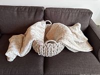 Two cream-colored plush blankets and a chunky knitted basket placed on a brown couch, showing texture and overall good condition of items.