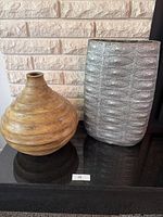 Two large decorative vases placed on a dark surface in front of a brick wall. The silver one on the right has a textured finish and is taller; the brown one on the left is rounded with distinct rings.