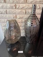 Two large decorative vases with differing shapes and finishes, placed on a black surface in front of a light brick wall.