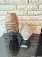 Photo of two decorative vases on a black surface against a white brick wall background, showing the taller ribbed vase and the shorter striped vase side by side.