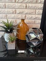 Photo showing two faux plants, brown ceramic vase, and silver geometric decor on black reflective surface in front of white brick wall.