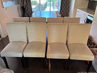 Front-facing view of eight eggshell white upholstered dining chairs arranged in two rows of four on a dark wood floor.