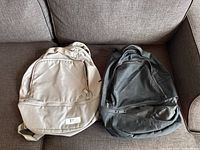 Two Lululemon backpacks side by side on a couch, one light gray and one black, showing overall shape and color.