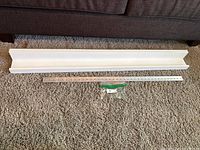 Top view of white Pottery Barn decor shelf against a carpeted floor. A metal mounting bracket and small bag of screws placed alongside for scale and installation.