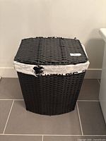Closed black wicker laundry basket against a white wall and grey tile floor showing slight wear on the fabric liner near the rim.