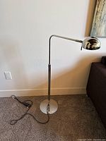 Full view of stainless steel standing lamp showing tall adjustable pole, curved arm, dome-shaped lamp head, power cord and foot switch on carpeted floor.