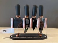 Frontal view of four black handled cheese knives with copper accents displayed in a clear acrylic and black base stand.