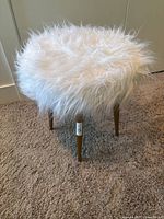 Front angle view of a round faux fur upholstered stool with wooden legs on carpeted floor