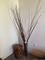 Wooden log stool and glass vase filled with tall decor sticks, viewed from a distance against a corner wall.