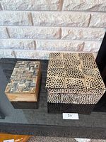 Photo showing two wooden boxes on a dark reflective surface against a white brick wall background. The smaller box has a mosaic stone top; the larger box has a beige and black patterned top.