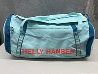 Side view of light blue Helly Hansen duffel bag showing 'HELLY HANSEN' text in large red letters.