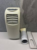 Front view of the portable air conditioner with vent hose and window adapter placed beside it.