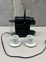 Black Nespresso coffee machine with two white espresso cups and saucers in front on a white surface against a gray wall