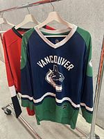 Vancouver Canucks hockey jersey on hanger showing front with team name and logo, and part of another jersey behind