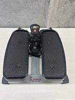Front view of the stepper showing two black textured foot pedals mounted on a silver metal frame with a central digital display screen and red control button, branded Sunny Health & Fitness.