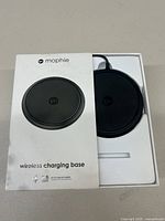 Top view of Mophie wireless charging base in original white box packaging. Shows black round charging surface with logo and power output detail.