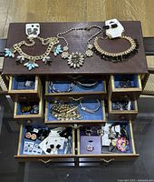 Top view of wooden jewelry cabinet with multiple drawers opened showing blue fabric lining and assortment of costume jewelry including necklaces with floral and bead designs, bracelets, earrings on cards, and pins.