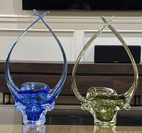 Both blown art glass freeform baskets shown side by side on a glass table, featuring one blue and one green basket with high arches.