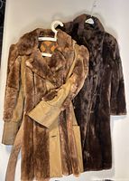 Two vintage sheared beaver fur coats hanging on hangers, showing different colors and leather trim on one coat.