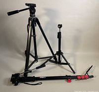 Three tripods displayed against a white backdrop: a larger Soligor T170 tripod, a smaller Cameron W-802 tripod, and an Ambico monopod with red clamps lying in front.
