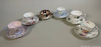 All six bone china teacup and saucer sets arranged in a row on a grey surface, showing different patterns and colors.